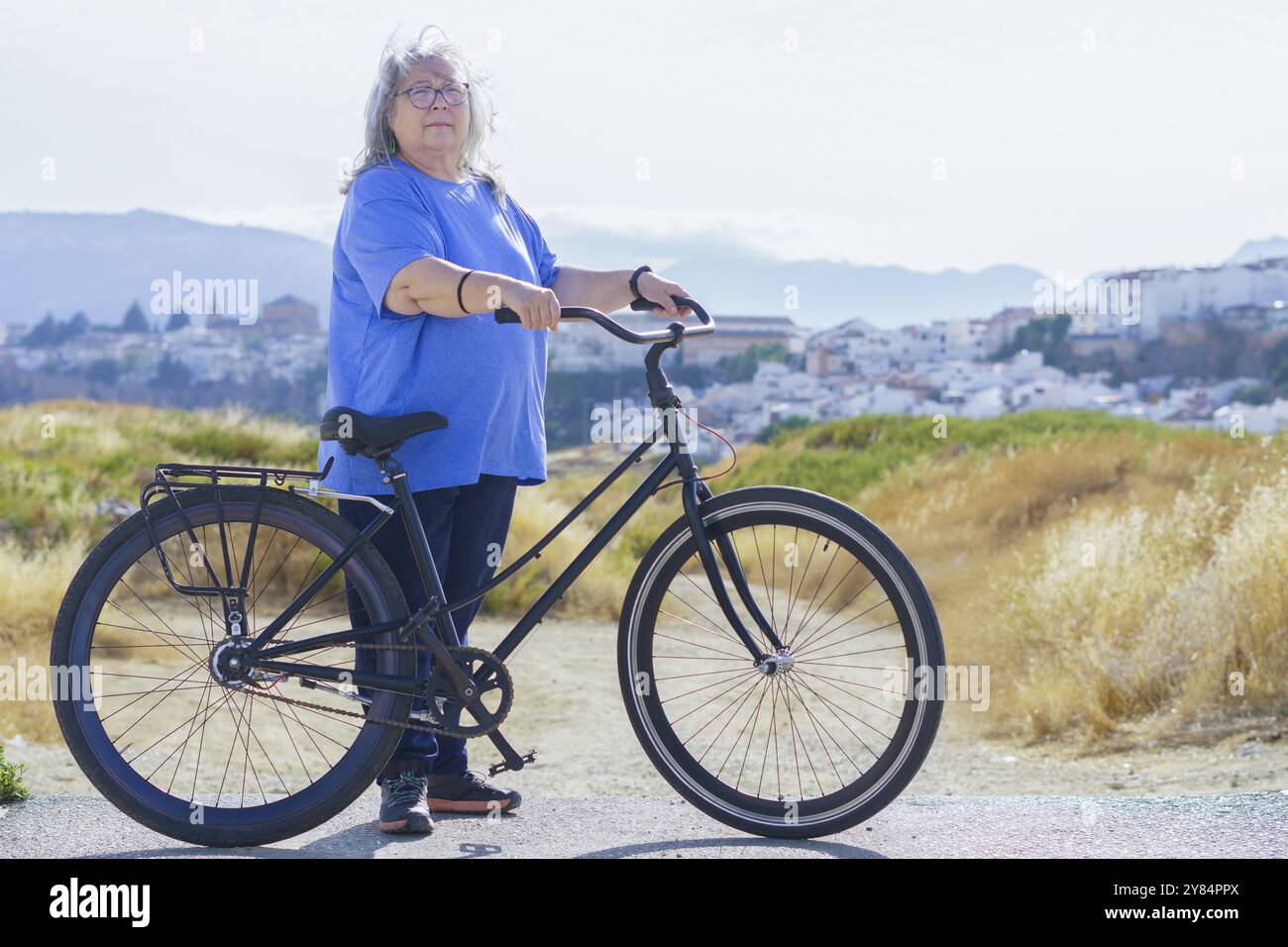 Femme âgée aux cheveux blancs en surpoids chevauchant un vélo de style rétro avec une ville en arrière-plan Banque D'Images