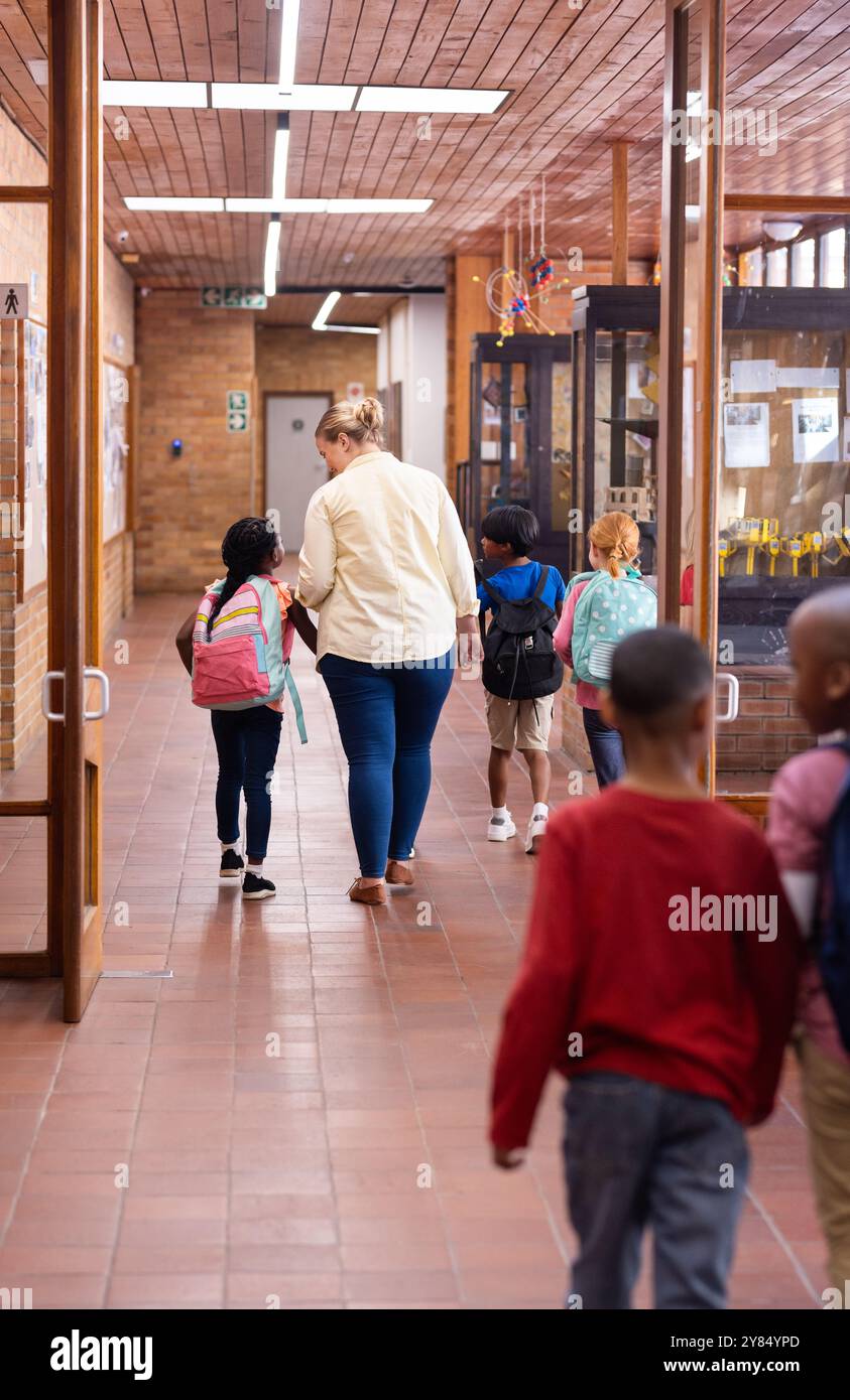 Marchant dans le couloir de l'école, enseignante guidant de jeunes élèves divers avec des sacs à dos Banque D'Images