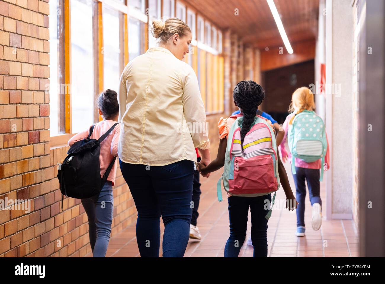 Marcher dans le couloir de l'école, enseignante tenant la main d'élèves divers avec sac à dos Banque D'Images