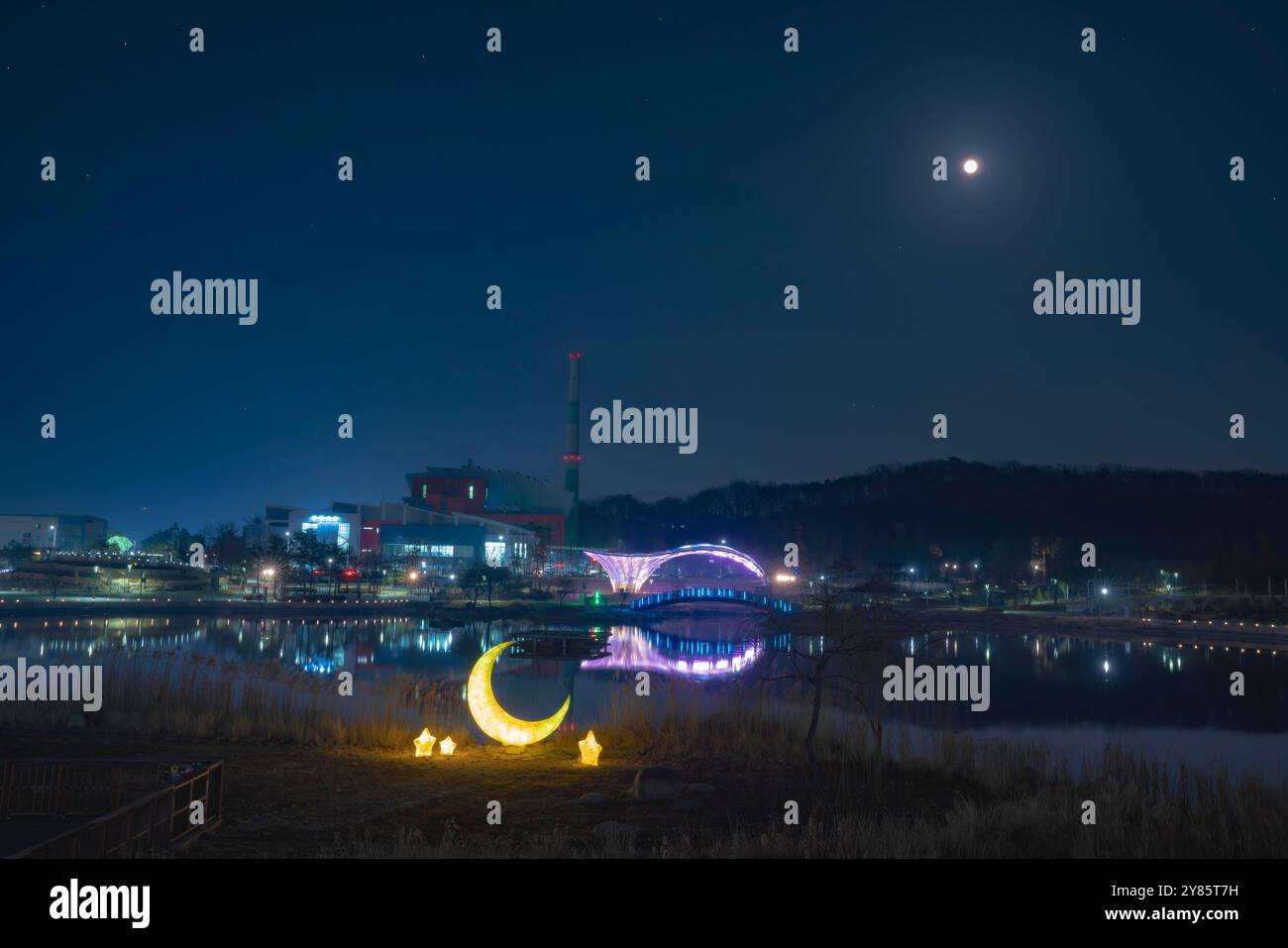 Nuit tranquille à Gimpo Lake Park avec la pleine lune. Le pont illuminé et la sculpture en croissant se reflètent sur l'eau calme, créant une scène paisible. Banque D'Images