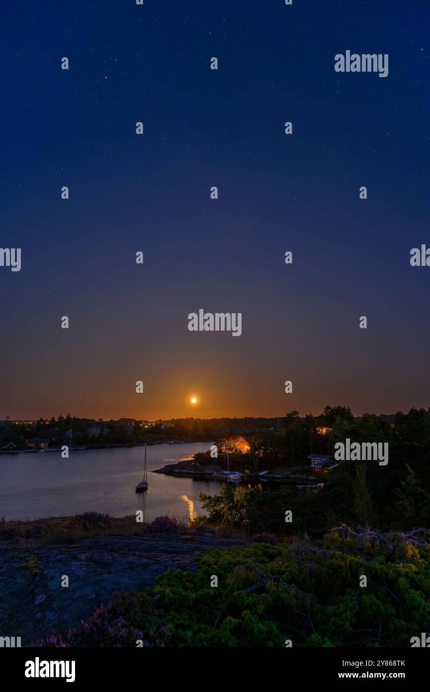 Lever de la lune et étoiles dans un ciel bleu clair de nuit sur les îles de l'archipel de Kragero avec voilier amarré et maisons d'été dans le comté de Telemark, en Norvège. Banque D'Images