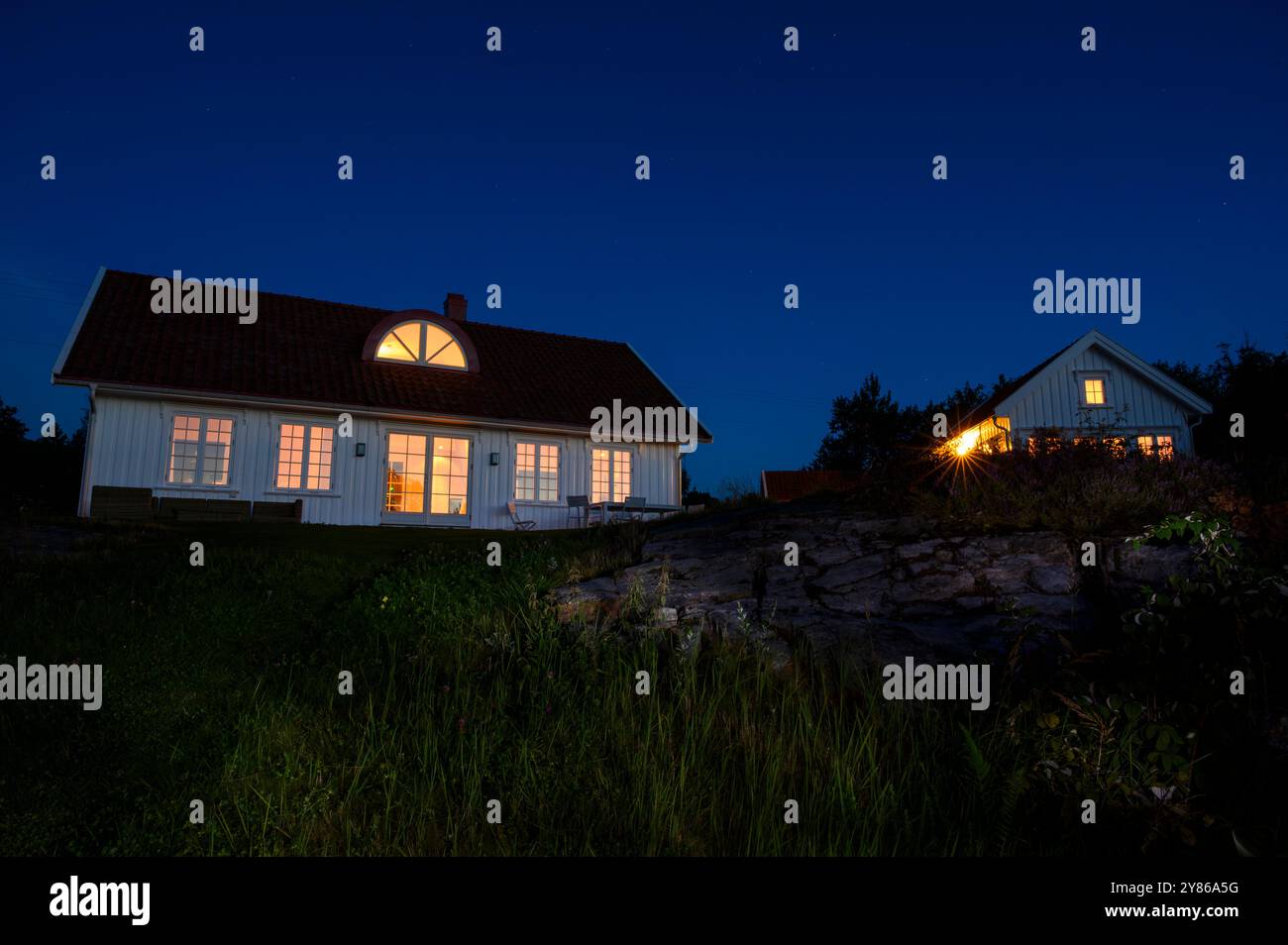 Maisons d'été la nuit avec la lumière ambiante brillante à travers les fenêtres contre un ciel bleu foncé et en partie éclairé au premier plan herbeux. Telemark, Norvège. Banque D'Images