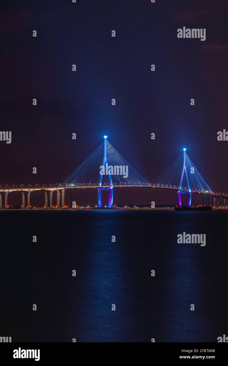 Une vue nocturne d'un pont magnifiquement illuminé, avec des lumières bleues et violettes reflétant sur l'eau calme, créant une atmosphère sereine. Banque D'Images