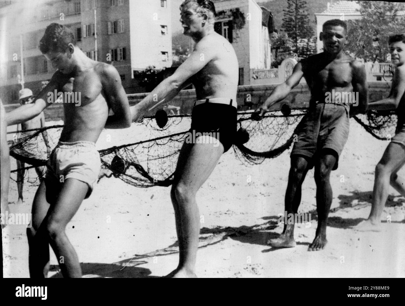 Eden donne un coup de main à Rio - Anthony Eden, (deuxième à partir de la gauche), ancien ministre britannique des Affaires étrangères, aide les pêcheurs à tirer leurs filets sur la plage de Rio de Janeiro, au Brésil, où il a passé de brèves vacances. Chaque matin et après-midi, un pêcheur local traîne le surf sur la plage pour trouver du poisson frais. 6 avril 1955. (Photo d'Associated Press photo). Banque D'Images