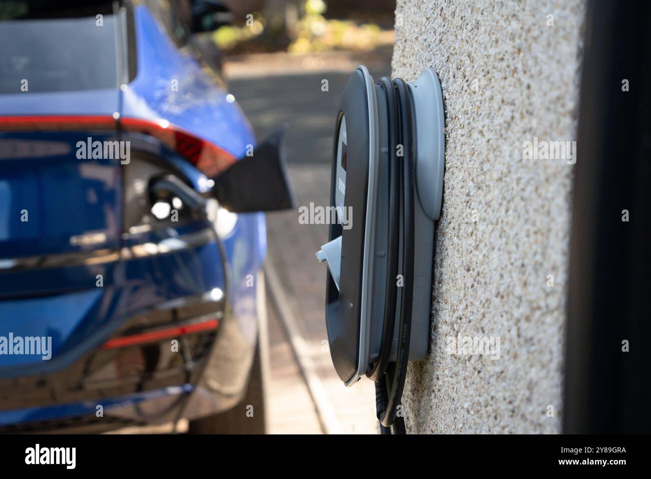 Une voiture électrique bleue se chargeant sur un chargeur domestique par une journée ensoleillée Banque D'Images