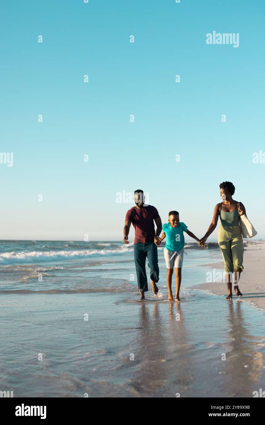 Garçon afro-américain ludique tenant père et mère les mains et courant dans la mer contre le ciel Banque D'Images