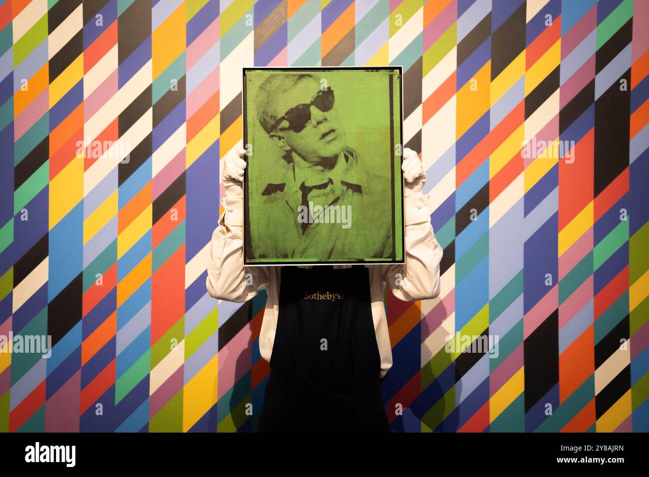 Un gestionnaire d'art tient l'Autoportrait d'Andy Warhol devant Gaillard 2 de Bridget Riley, lors d'un aperçu de la vente d'œuvres d'art Sotheby's de 200 millions de livres sterling, qui comprend des œuvres de Hockney, Warhol et Picasso. Date de la photo : vendredi 4 octobre 2024. Banque D'Images