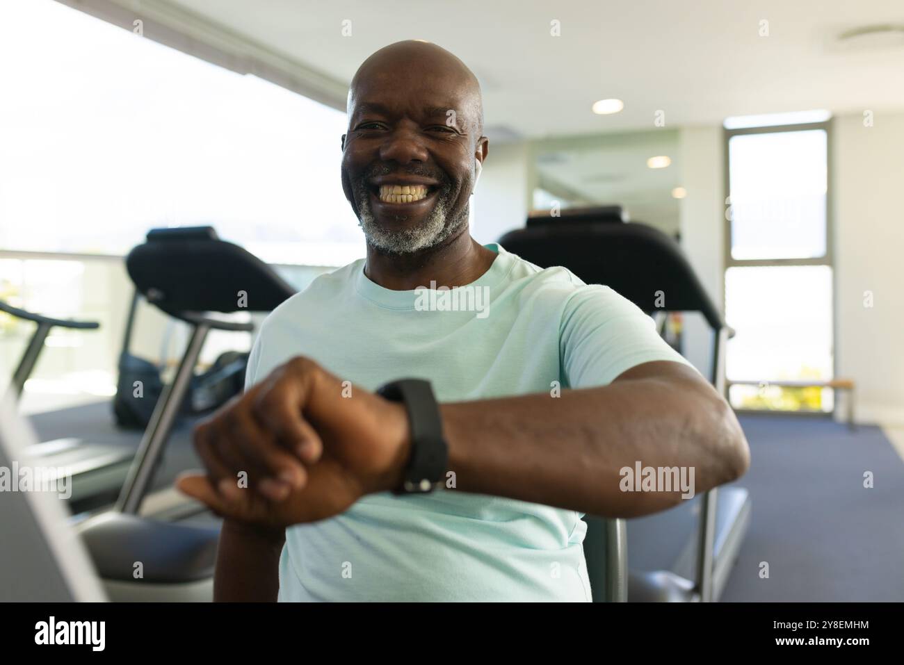 Homme senior afro-américain utilisant une smartwatch tout en étant assis sur le vélo stationnaire à la salle de gym Banque D'Images