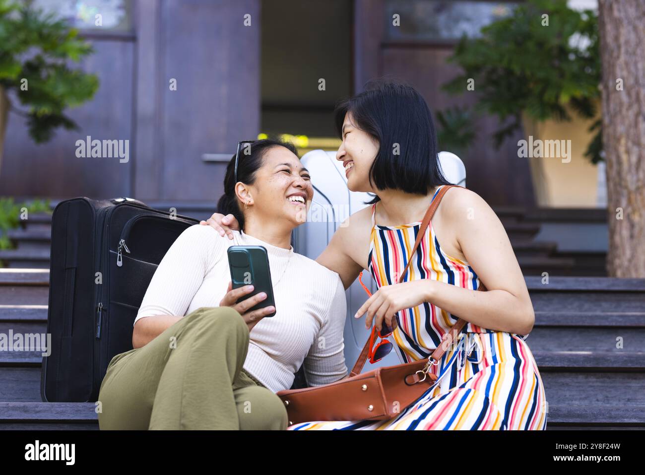 Heureuses amies asiatiques avec des bagages parlant avec smartphone dans le jardin à la maison Banque D'Images