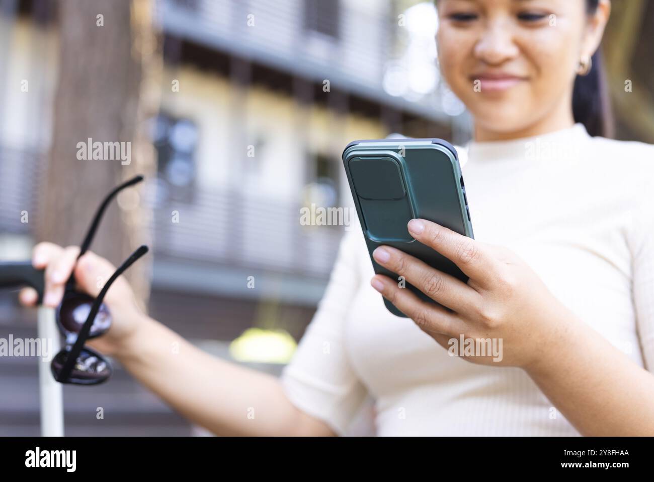 Femme asiatique heureuse tenant des lunettes de soleil et utilisant un smartphone dans le jardin à la maison Banque D'Images