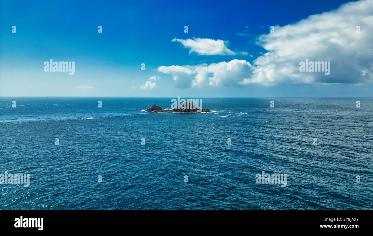 Une vue sereine d'une petite île rocheuse entourée d'eaux calmes bleues de l'océan sous un ciel clair avec des nuages blancs moelleux. La lumière du soleil se reflète sur le W. Banque D'Images