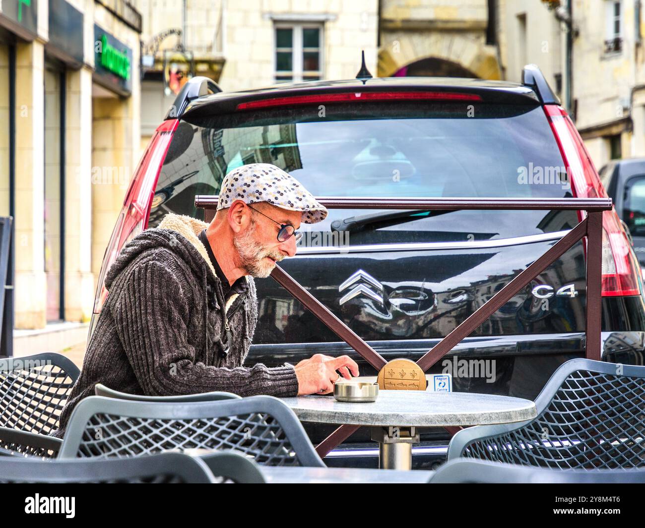 Homme en bonnet de tissu assis à la table de café extérieure vérifier smartphone - Loches, Indre-et-Loire (37), France. Banque D'Images