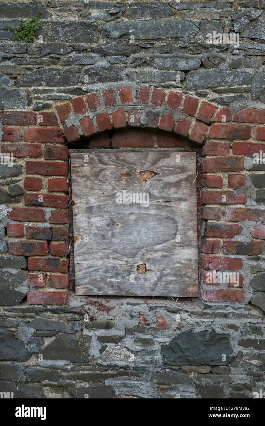Le mur d'un bâtiment industriel abandonné présente des pierres et des briques en ruine, avec des fenêtres en bois, évoquant un sentiment de délabrement et de négligence. Photo ta Banque D'Images