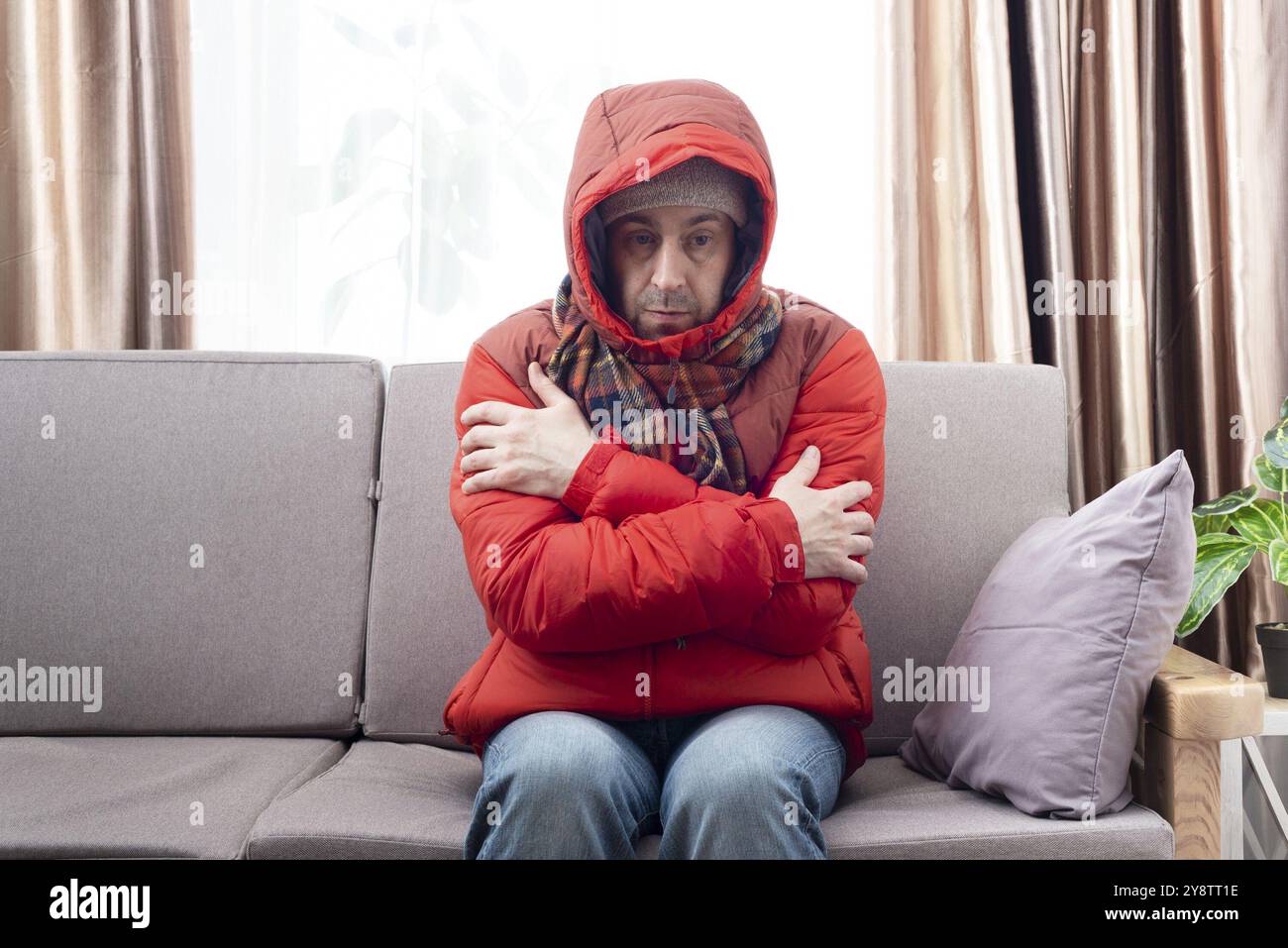 Modèle masculin moyen âge en parka rouge et écharpe à carreaux devenant froide assis sur le canapé dans le salon froid Banque D'Images