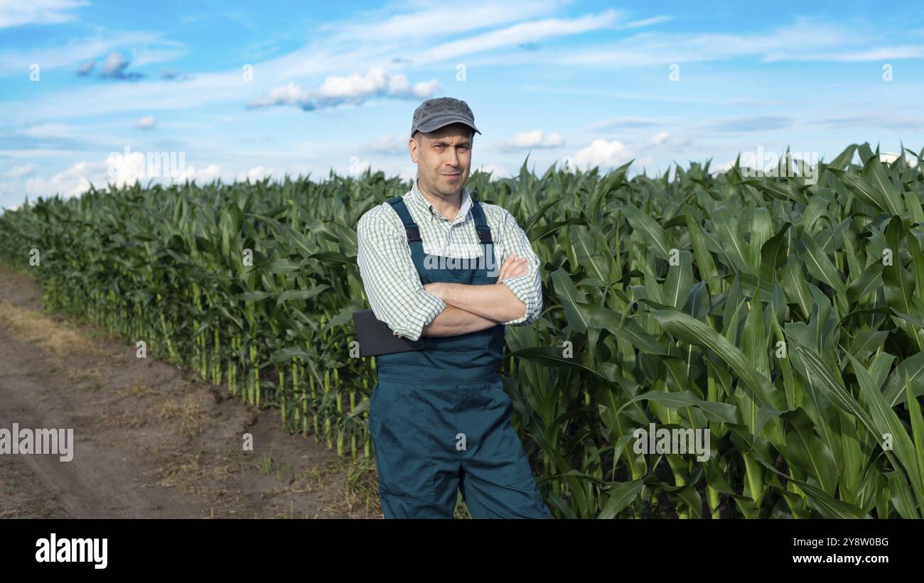 Un homme d'âge moyen caucasien satisfait confiant travailleur agricole dans des combinaisons avec des bras croisés au champ de maïs Banque D'Images