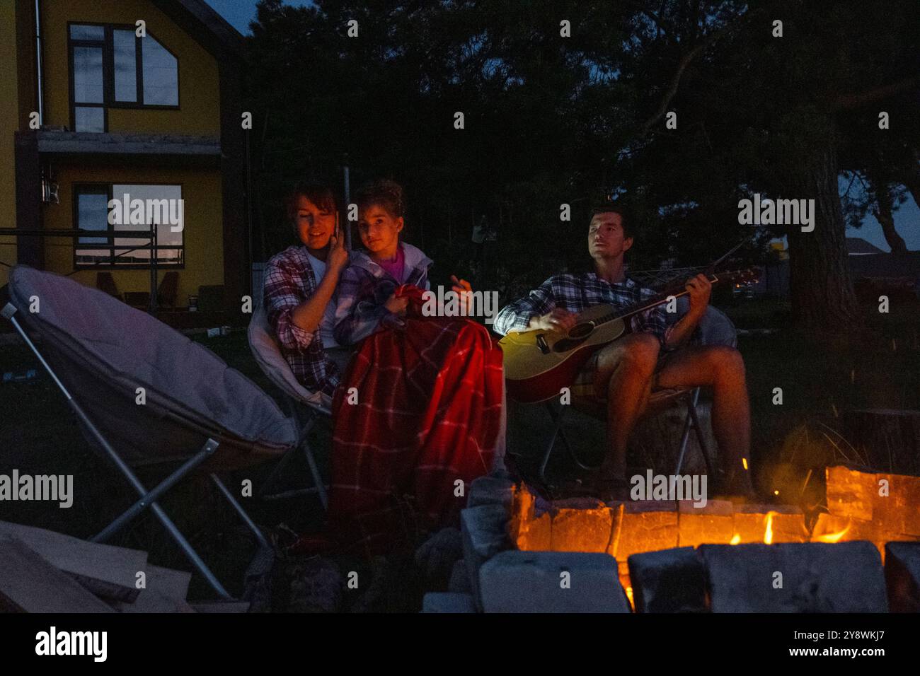 La famille est assise autour du feu de camp, un homme joue de la guitare, une femme écoute et chante, ma fille s'est enveloppée dans une couverture. Somme famille Banque D'Images