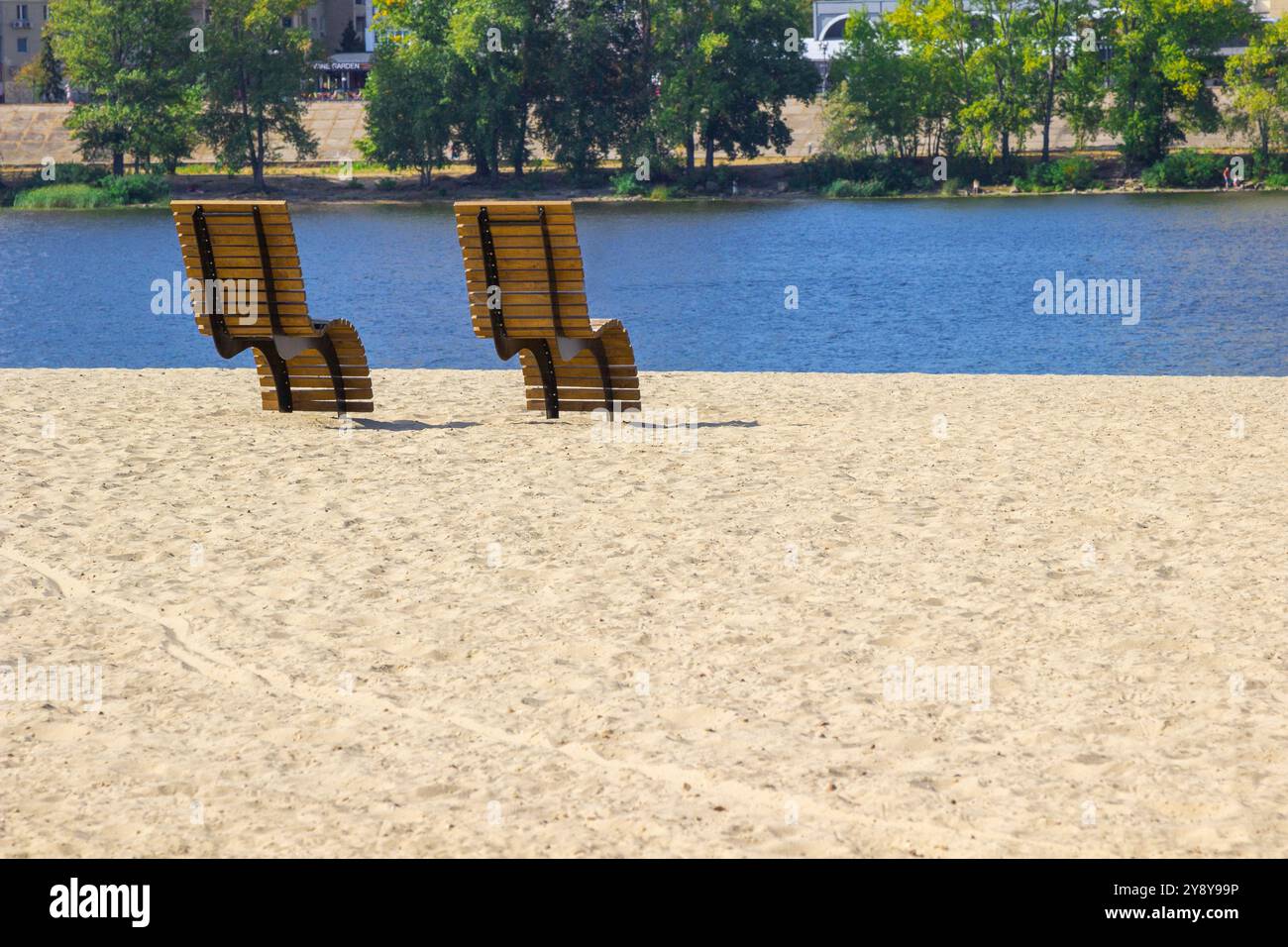 Deux chaises longues en bois sur une plage de sable. Rivière bleue. Arbres de l'autre côté de la rivière. Banque D'Images