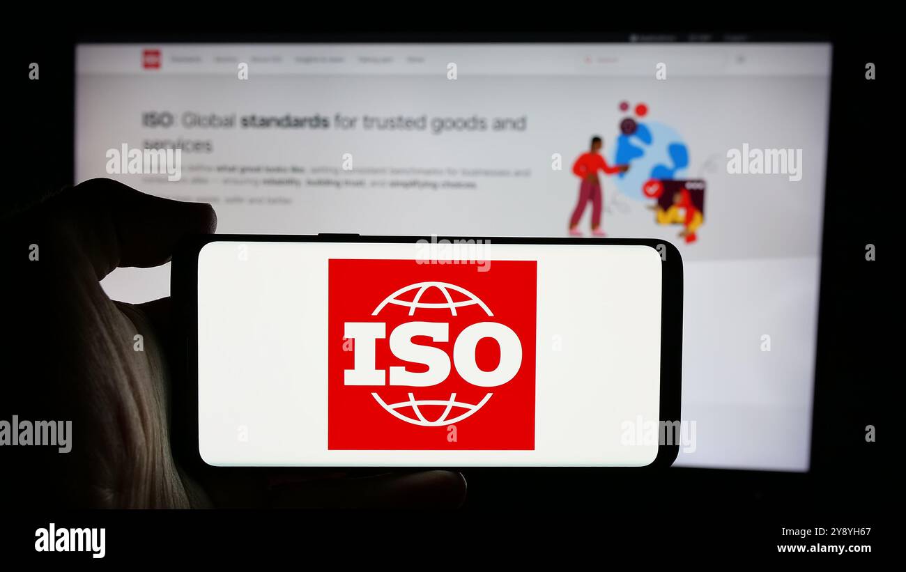 Personne tenant un téléphone portable avec le logo de l'Organisation internationale de normalisation (ISO) sur l'écran devant la page Web. Concentrez-vous sur l'affichage du téléphone. Banque D'Images
