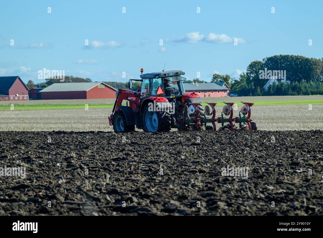 Une journée brillante à la campagne alors qu'un tracteur laboure efficacement le champ, retournant le sol tandis que des granges parsèment l'horizon, mettant en valeur l'activité agricole Banque D'Images