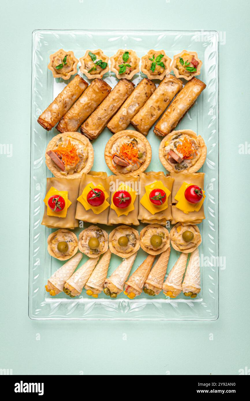 Un élégant plateau de collation avec des rouleaux de printemps, des rouleaux de lasagne recouverts de tomates cerises, des mini tartes et des hors-d'œuvre salés en forme de cône sur un plat en verre. Banque D'Images