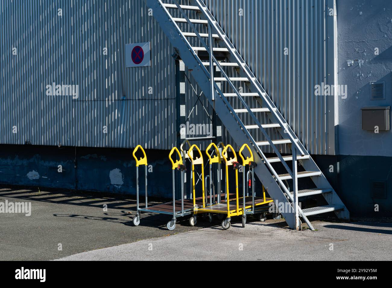 Plusieurs chariots jaunes sont soigneusement disposés à côté d'un escalier métallique à l'extérieur d'une installation de stockage. La lumière du soleil projette des ombres, créant un contraste visuel saisissant sur le sol. Banque D'Images