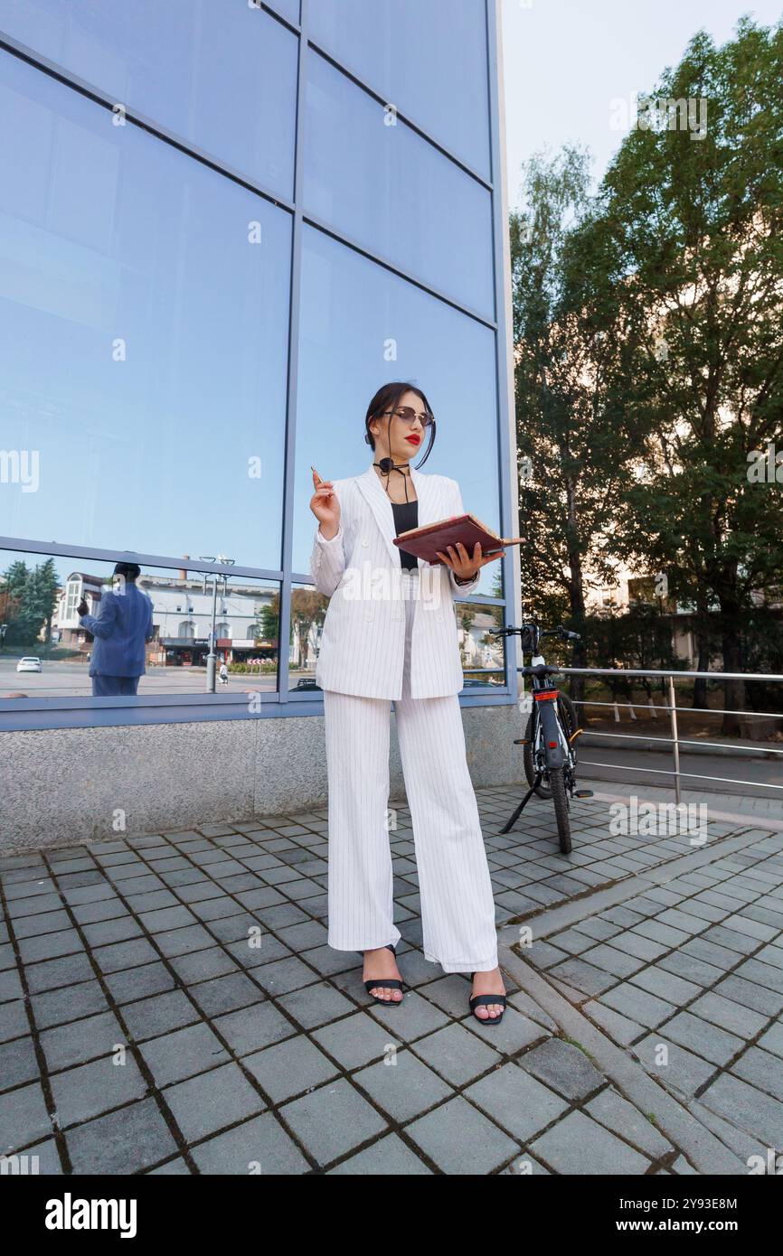 Femme d'affaires élégante en costume rayé tenant un cahier près de l'architecture moderne. Banque D'Images