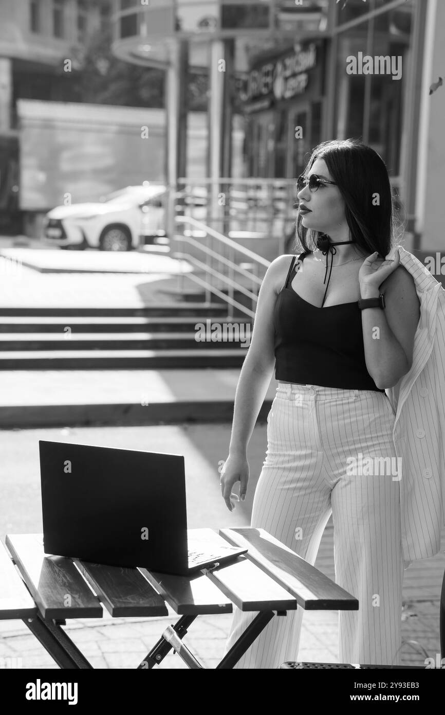 Une femme d'affaires élégante en lunettes de soleil posant en toute confiance à l'extérieur avec un ordinateur portable et un Blazer chic. Banque D'Images