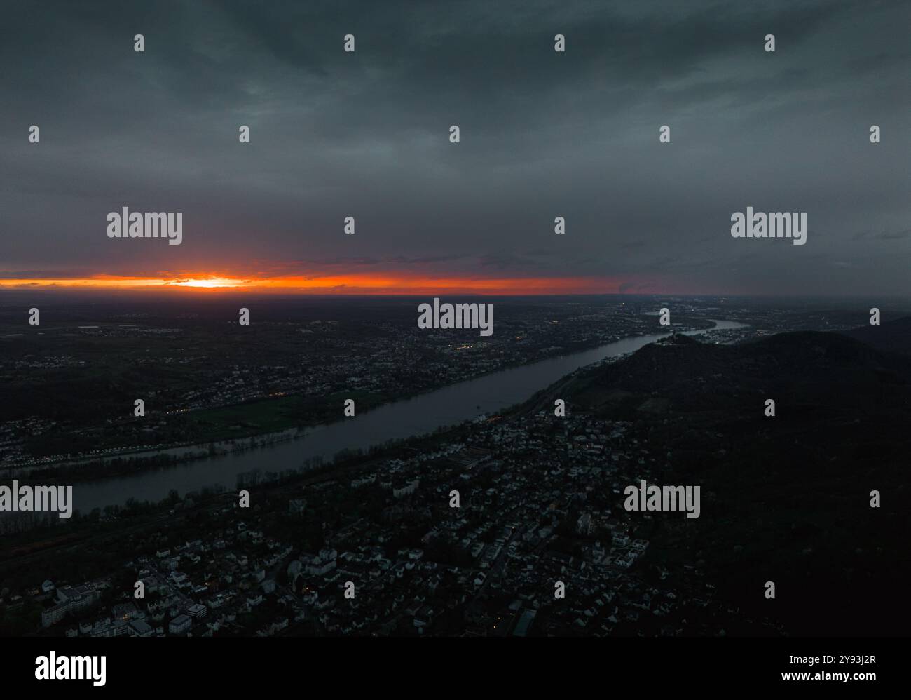 Coucher de soleil spectaculaire sur un paysage fluvial avec un horizon orange vibrant, des nuages sombres et un paysage urbain tentaculaire en contrebas, créant un contraste captivant. Banque D'Images