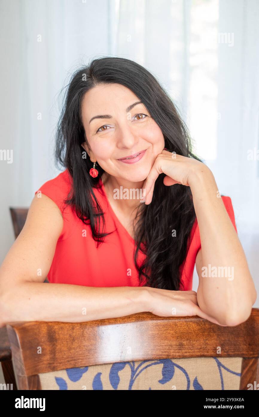 Une femme dans une robe rouge décontractée sourit tout en étant assise à une table, créant une atmosphère lumineuse et joyeuse, concept d'expression amicale Banque D'Images