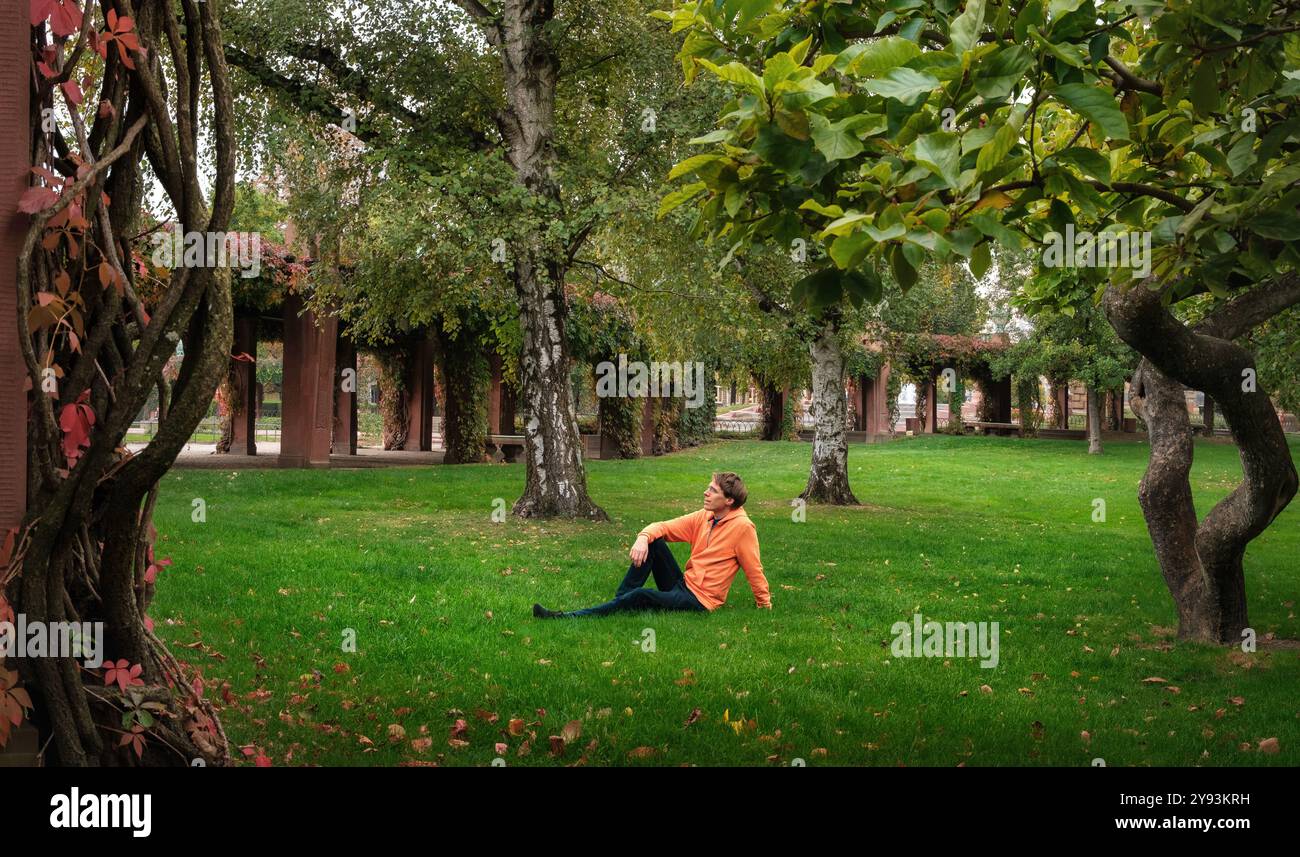 Homme détendu assis seul sur l'herbe luxuriante dans un parc verdoyant pittoresque, encadré par des troncs rongés et des plantes Banque D'Images