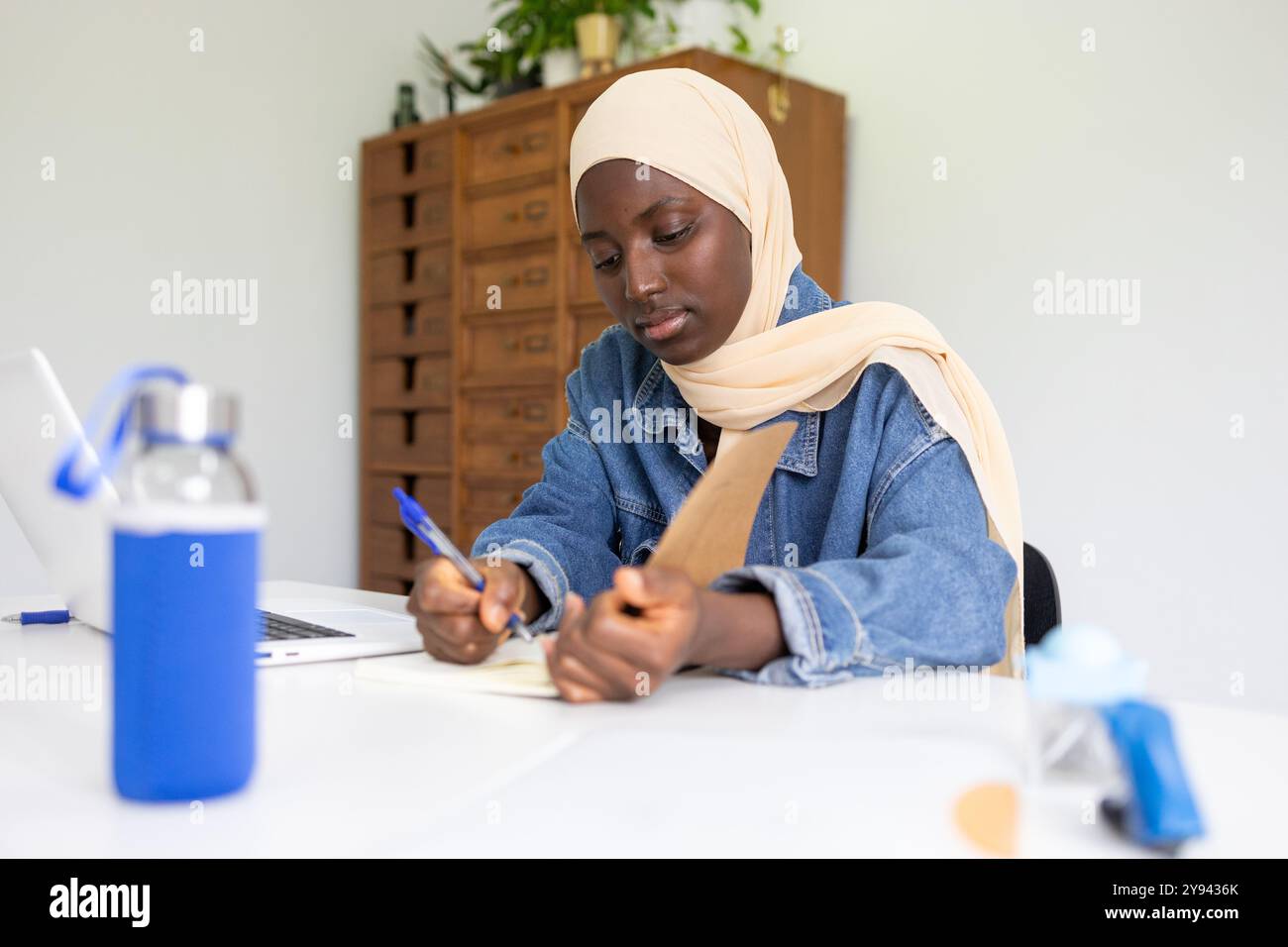 Une femme noire concentrée médecin portant un hijab est assise à son bureau, prenant diligemment des notes d'un livre elle est entourée d'objets, y compris un ordinateur portable et Banque D'Images