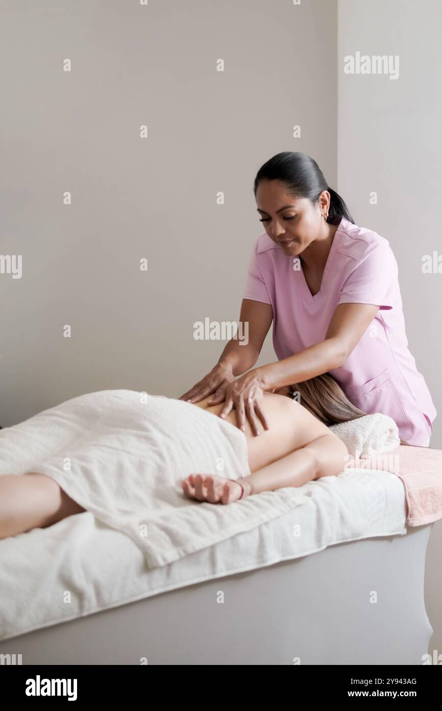 Une thérapeute féminine dans un uniforme rose effectue un massage du dos apaisant sur une cliente couchée face vers le bas sur une table de massage à la maison. Le thérapeute l'est Banque D'Images