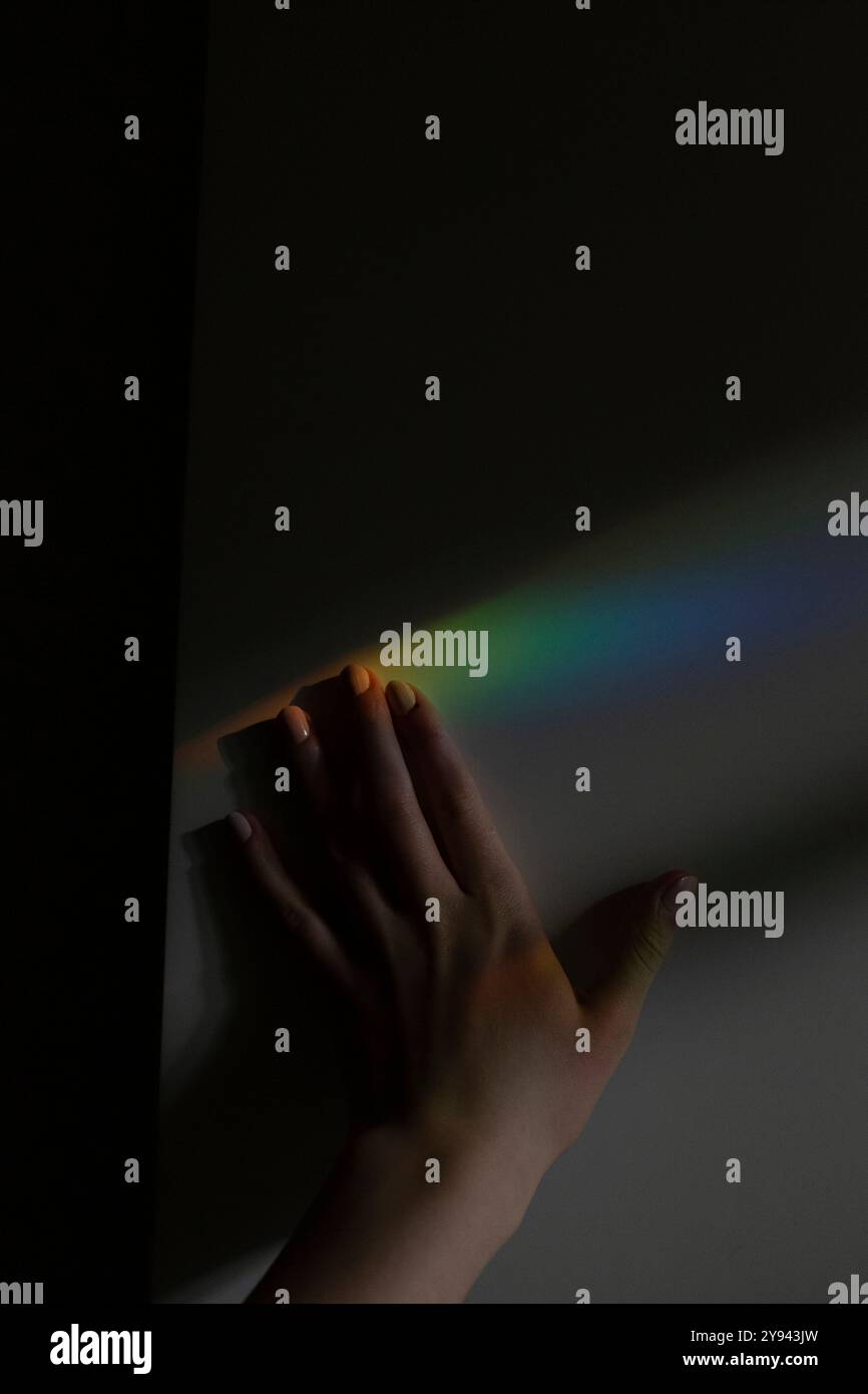 Image artistique d'une main s'approchant d'une douce lumière arc-en-ciel réfléchie sur un mur sombre la lumière et l'ombre contrastées créent un éthérée, minimaliste Banque D'Images