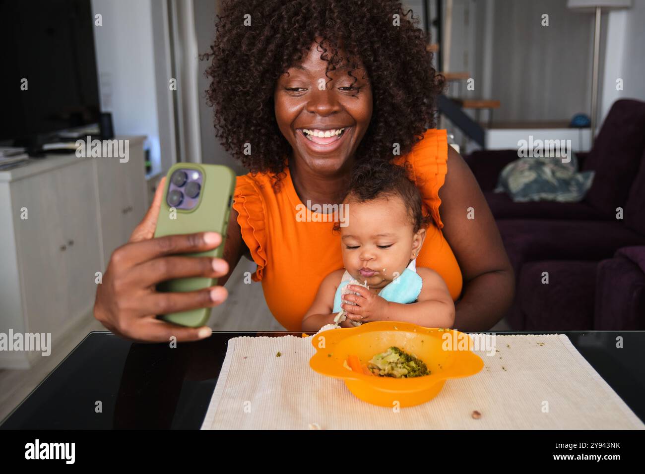 Une femme africaine prend joyeusement un selfie avec sa petite fille, qui mange dans un bol, capturant un moment joyeux et familial à la maison Banque D'Images