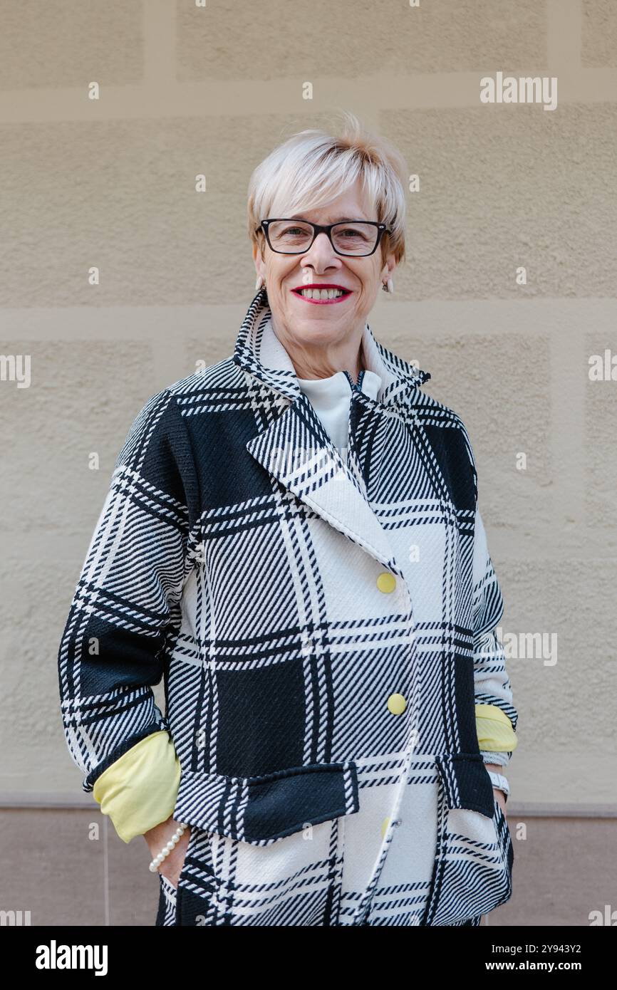 Une femme âgée optimiste aux cheveux blonds et aux lunettes, portant un manteau à carreaux noir et blanc à la mode, sourit chaleureusement contre un mur beige doux Banque D'Images