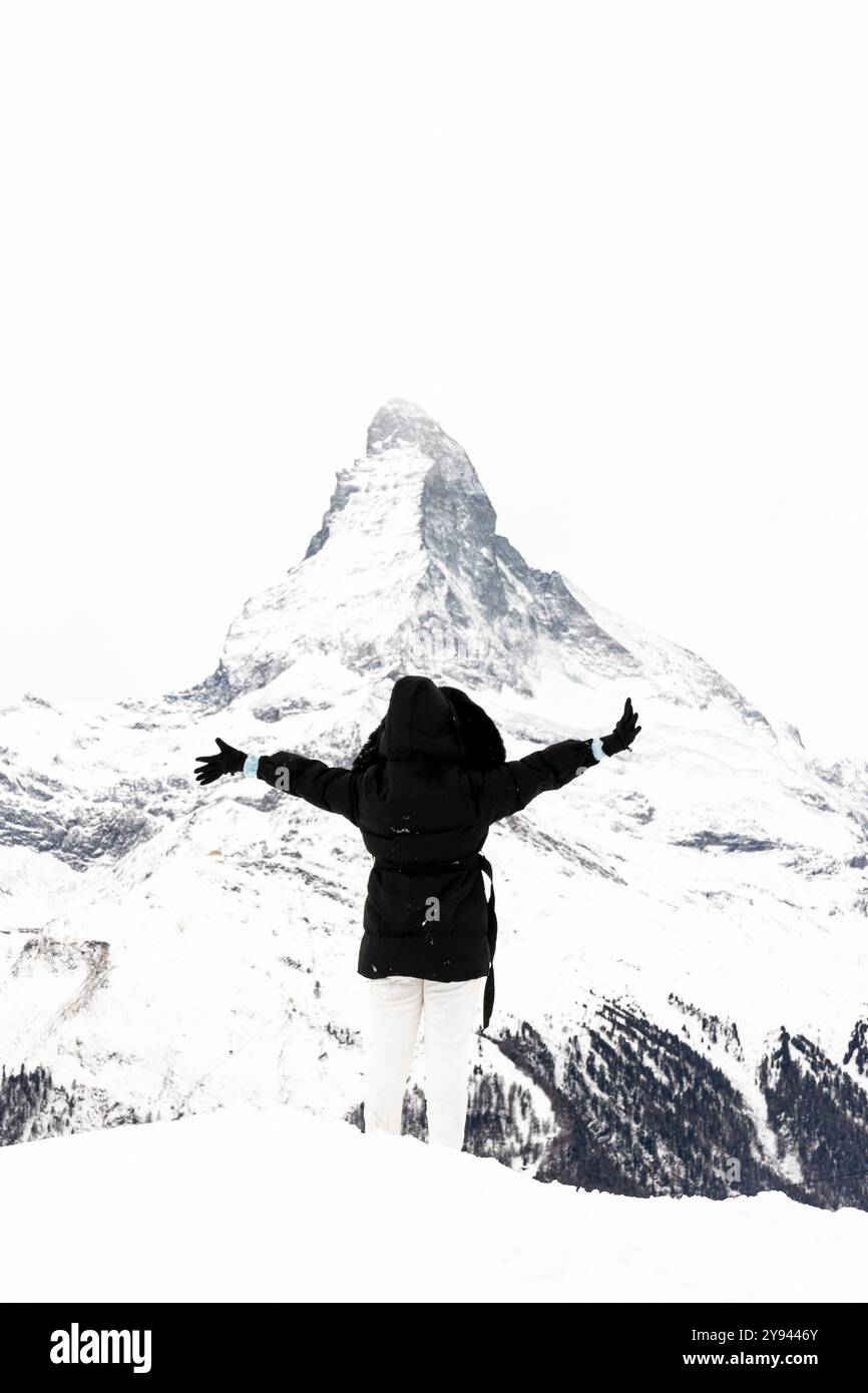 Vue arrière d'une personne méconnaissable debout dans la neige, les bras écartés, face à un majestueux sommet de montagne enneigé Matterhorn sur le fond. Banque D'Images