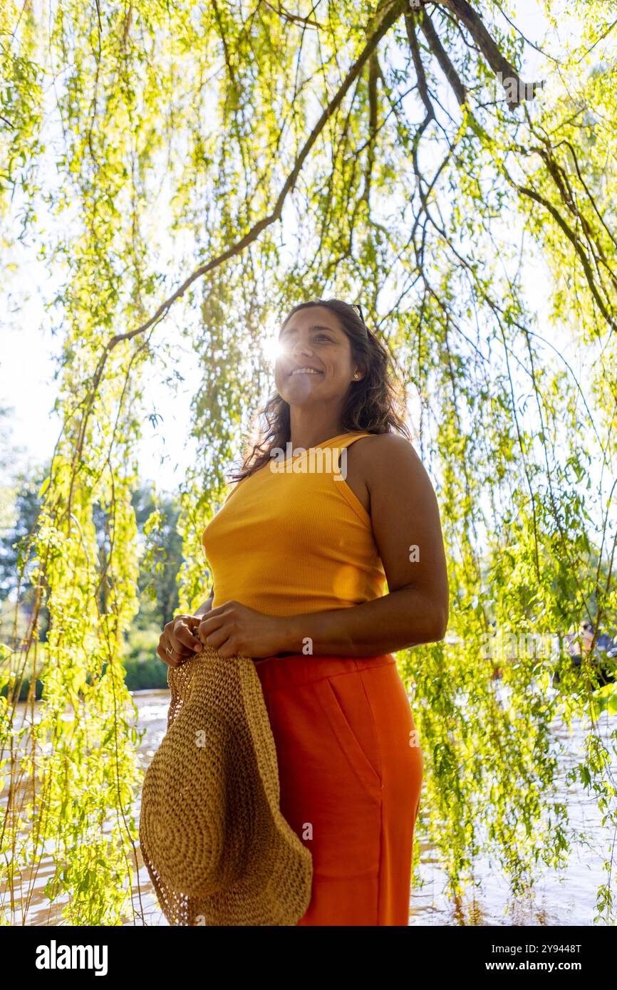 Une femme joyeuse dans un haut jaune et une jupe orange se tient sous les saules lors d'une journée d'automne ensoleillée à Amsterdam, exsudant bonheur et détente Banque D'Images