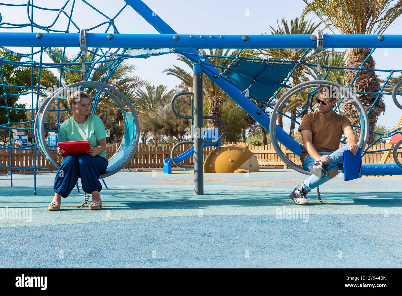 Un couple aime les balançoires dans un parc de plage assis sur des balançoires circulaires séparées, ils tiennent des tablettes, se livrant à leurs propres activités whil Banque D'Images