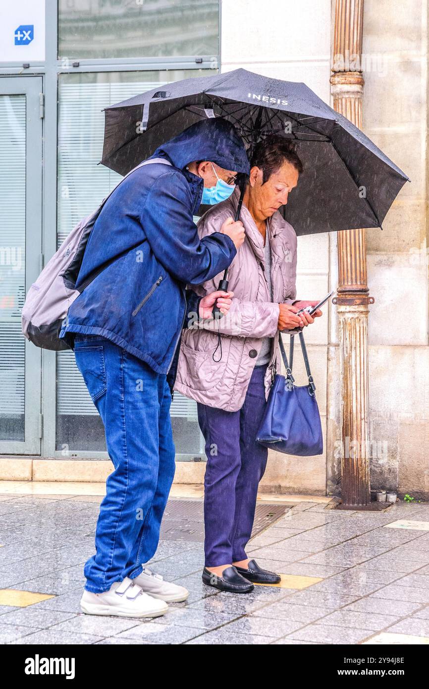 Couple oriental âgé sous parapluie sous une pluie battante regardant un téléphone portable - Tours, Indre-et-Loire (37), France. Banque D'Images