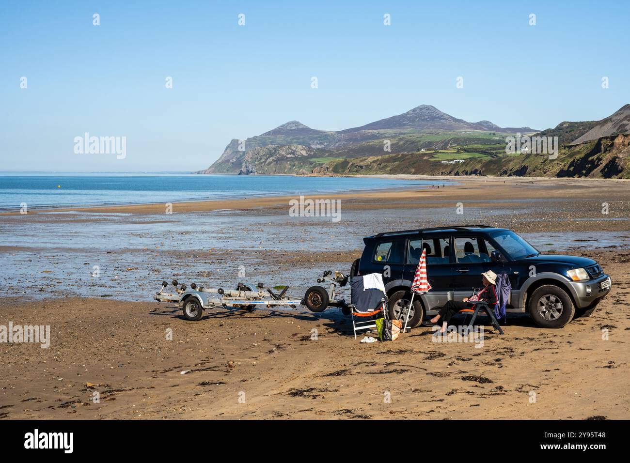 Une femme se détend dans un transat à côté d'un SUV sur la plage de Nefyn sur la péninsule de Llŷn au nord du pays de Galles, avec la colline Yr Eifl en arrière-plan. Banque D'Images