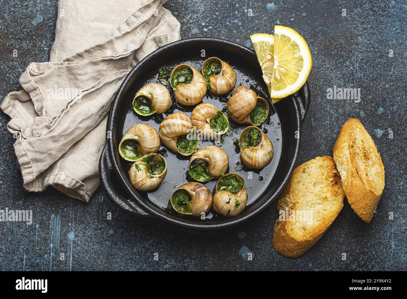 Escargots d'Escargotes de Bourgogne au beurre d'ail et au persil dans une casserole en fonte noire avec des tranches de citron et de baguette grillée sur fond de pierre rustique Banque D'Images