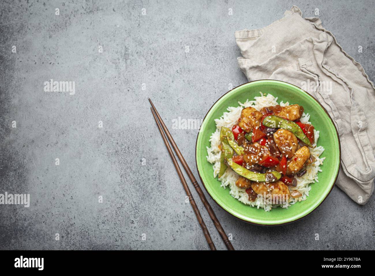 Poulet collant aigre-doux asiatique avec légumes sautés et riz dans un bol en céramique avec baguettes vue de dessus sur fond de pierre rustique gris, tradit Banque D'Images