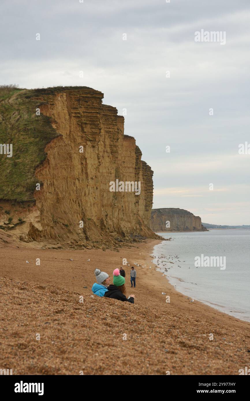 Un couple assis sur la plage portant des chapeaux bouloches en laine colorés. West Bay et les falaises de Golden Cap. Banque D'Images