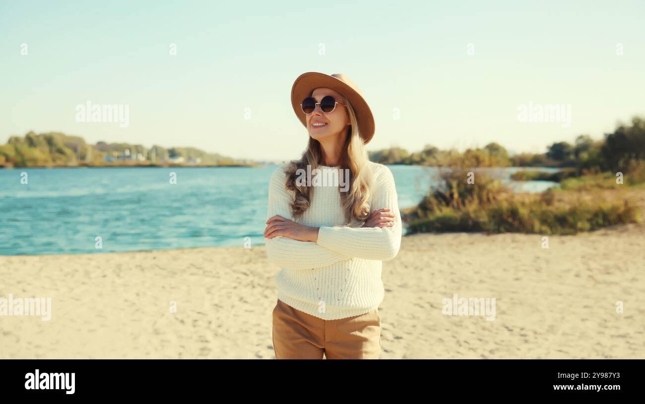 Femme mature élégante sur la plage côtière, modèle féminin d'âge moyen à la mode posant à l'extérieur, portant un pull, un chapeau et des lunettes, debout avec le bras croisé Banque D'Images