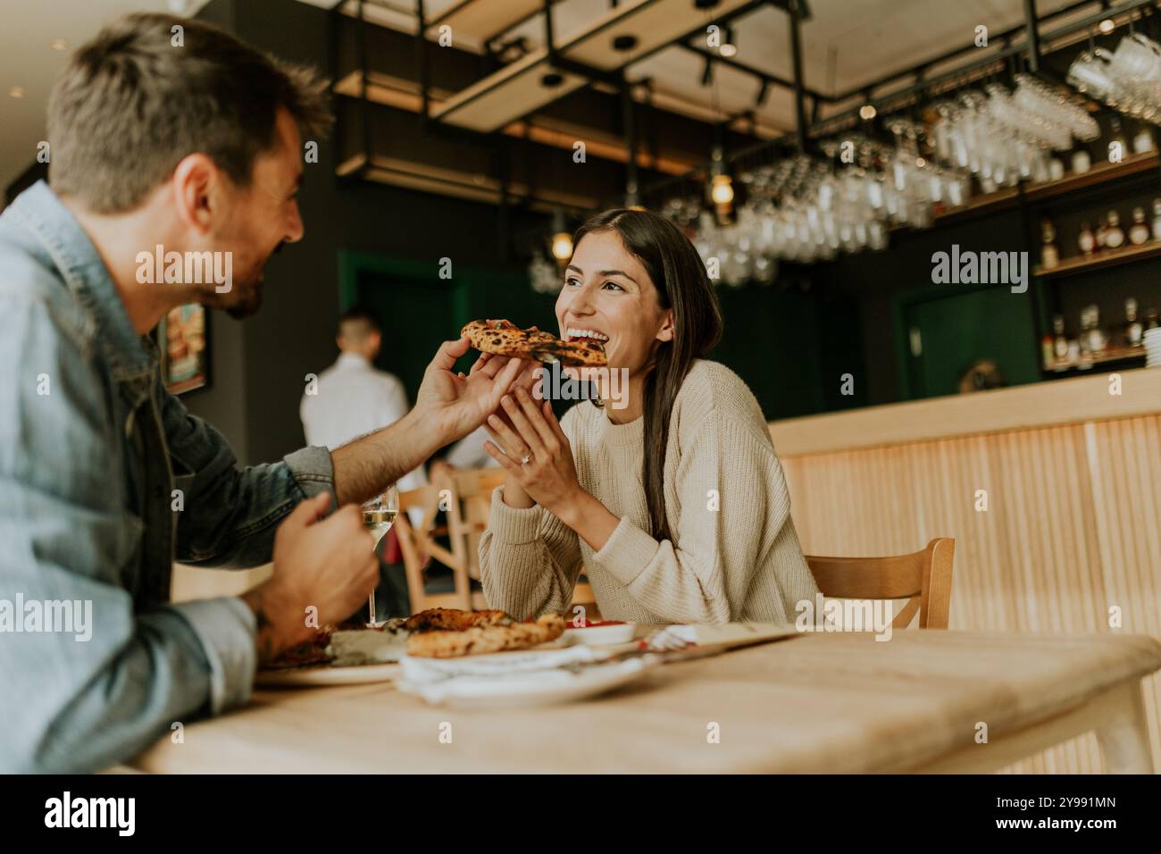 Un couple joyeux partage un moment dans un restaurant rustique, savourant des tranches de pizza tout en riant ensemble et en profitant de l'atmosphère vibrante autour de th Banque D'Images