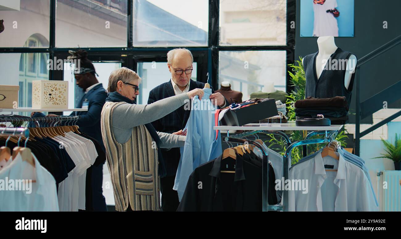 Les clients âgés analysent des chemises élégantes sur des cintres dans un magasin de détail, regardant une nouvelle ligne de vêtements sur l'affichage à la boutique. Couple senior à la recherche de vêtements formels, style de vie chic. Caméra B. Banque D'Images