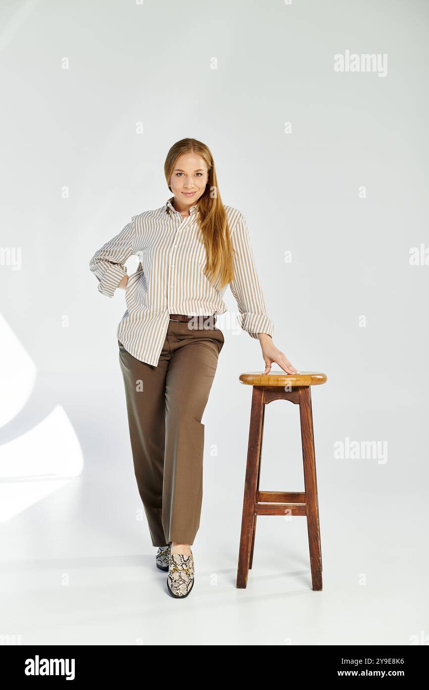 Une femme aux cheveux longs dans une chemise rayée pose à côté d'un tabouret en bois sur un fond gris neutre. Banque D'Images