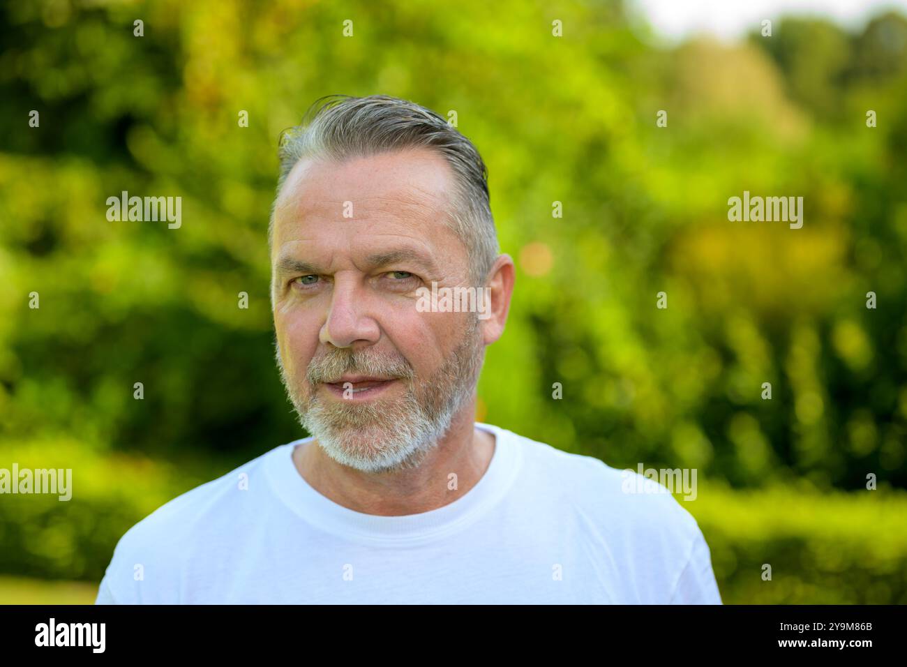 Un homme mature avec des cheveux gris et une barbe, portant un T-shirt blanc, se tient à l'extérieur avec un fond vert éclatant et flou. Banque D'Images