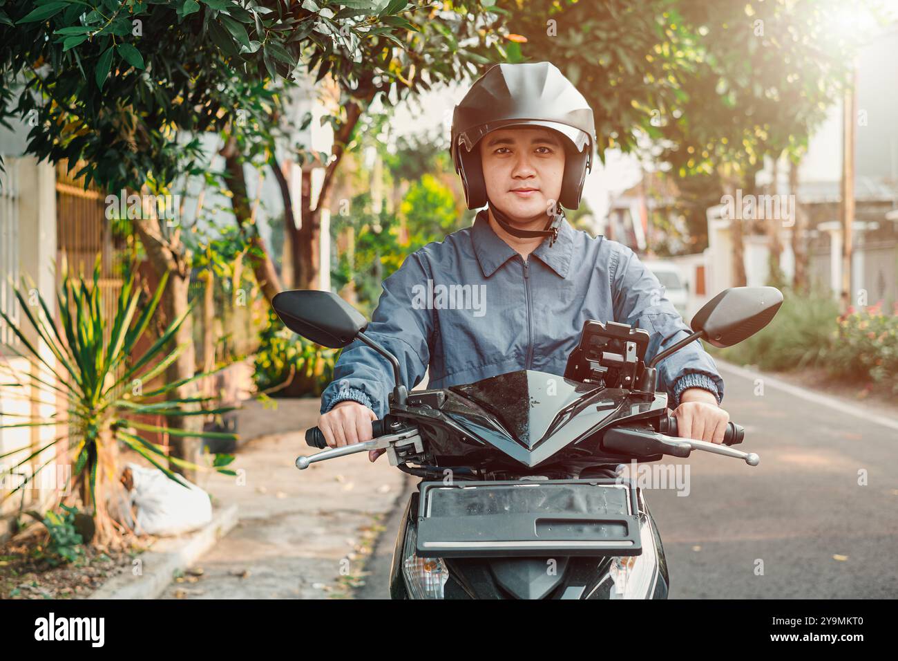 Un homme asiatique avec une veste bleue monte un scooter automatique ou une moto avec un support de téléphone intégré et regarde l'appareil photo pendant le coucher du soleil en t Banque D'Images