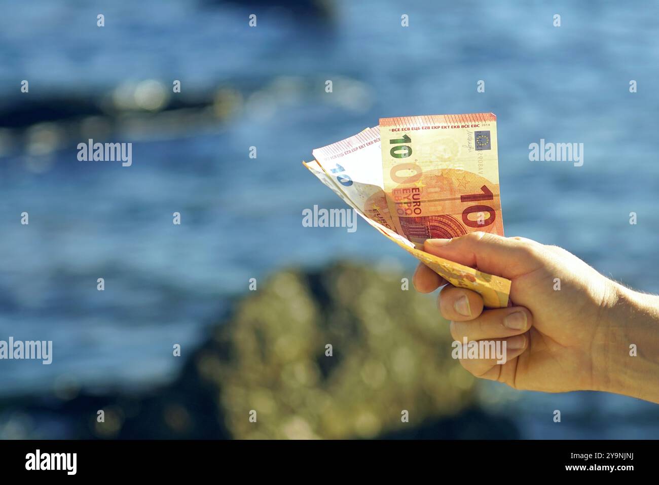 Dépenses pendant les vacances. Une photo en gros plan de la main d'un homme avec 70 euros (deux billets de 10 euros et un billet de 50 euros) sur fond de mer. Banque D'Images